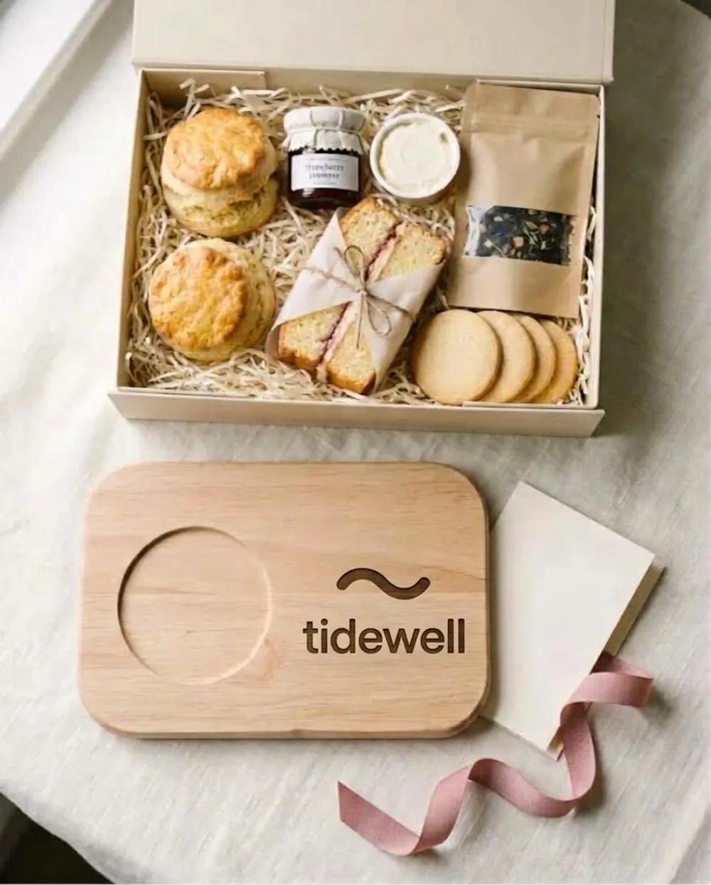 An afternoon tea box with scones, jam, cake, tea and biscuits, and a wooden board engraved with a logo