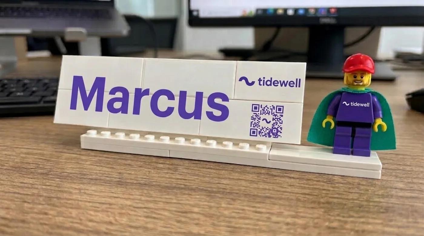 A white Lego name plaque printed with the name Marcus and a QR code, beside a minifig in a teal cape